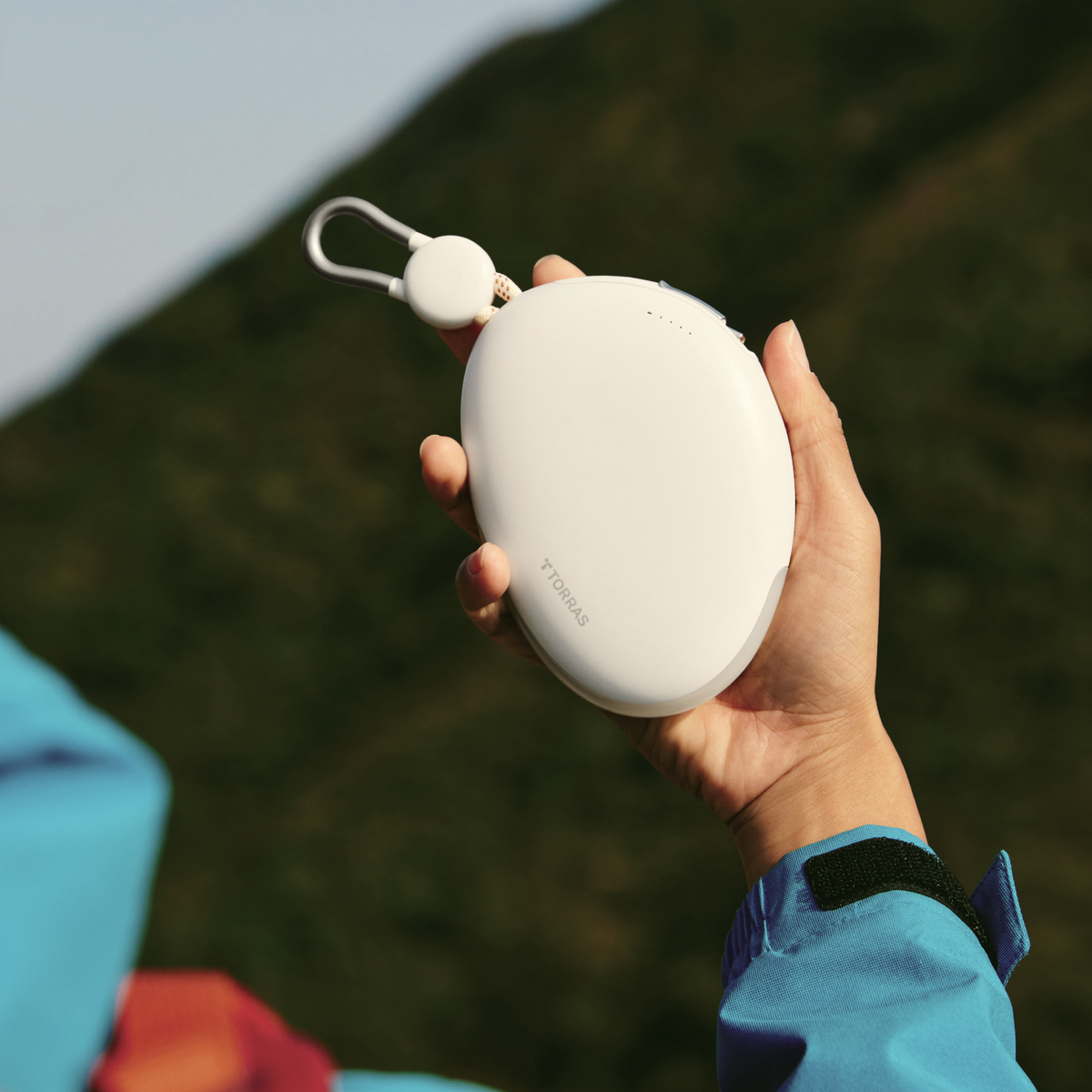 White TORRAS FlexLine Pebble Power Bank 10k mAh with lanyard and carabiner clip, held outdoors.